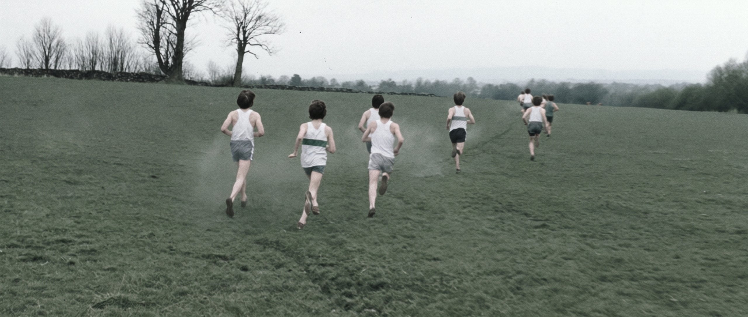 Cross-country runners crossing a misty green field, seen from behind — grainy 35mm documentary film still